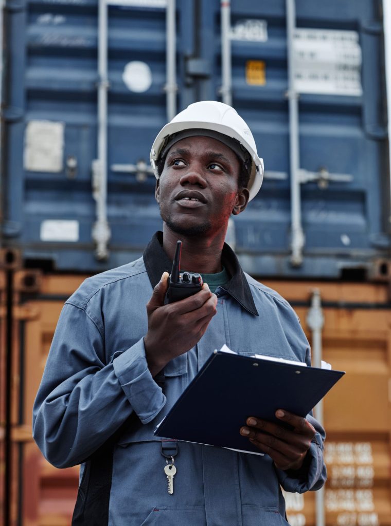 Vertical waist up portrait of black male worker wearing hardhat in shipping docks against containers